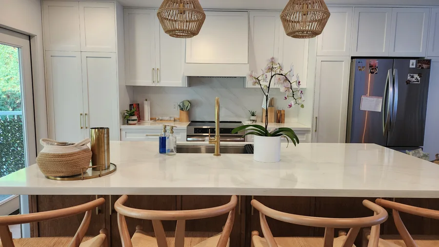 White kitchen with a broad pale island, timber stools and woven pendant lights.